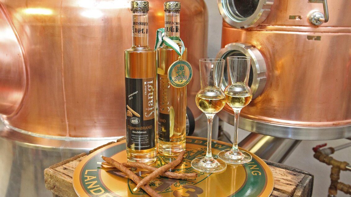 Zwei Flaschen Likör stehen auf einem dekorativen Teller neben Gläsern und Zimtstangen. Im Hintergrund sind kupferne Destillationsgeräte zu sehen. | © Obsthof Stangl