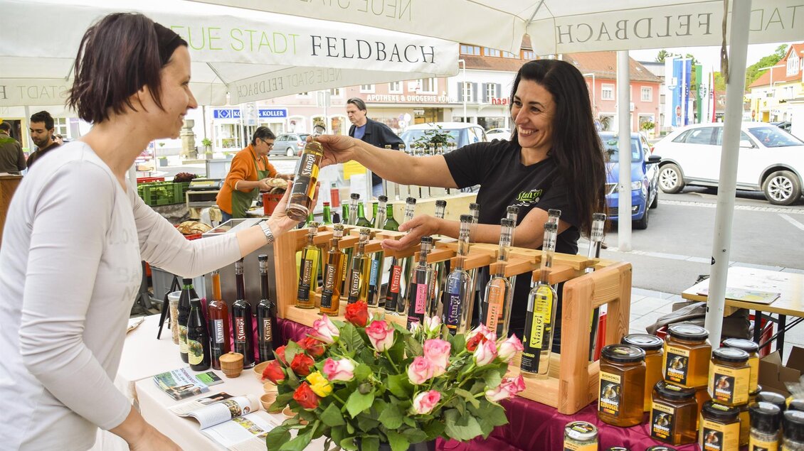Ein lebhafter Marktstand mit zwei Frauen, die miteinander sprechen. Es gibt verschiedene Flaschen und Produkte sowie Blumen auf dem Tisch. | © Thermen- & Vulkanland Steiermark