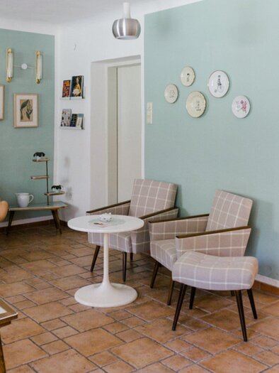 A stylishly furnished living room with soft colors and retro furniture. Pictures and plates hang on the wall, while a shelf with products can be seen in the background. | © Omas Teekanne