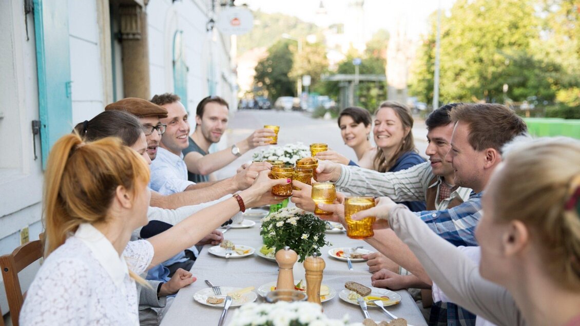 Eine Gruppe von Freunden erhebt die Gläser beim Essen im Freien. Der Tisch ist festlich gedeckt und es gibt Blumenmitten. | © Omas Teekanne - Svetlana Gombats Photography