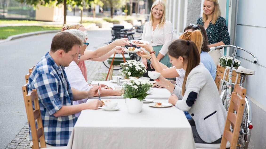 Eine Gruppe von Menschen sitzt an einem Tisch im Freien und genießt eine Mahlzeit. Alle stoßen mit ihren Tassen an und freuen sich über die gemeinsame Zeit. | © Omas Teekanne - Svetlana Gombats Photography