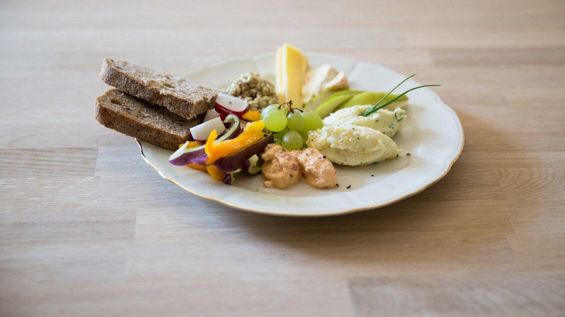Ein Teller mit verschiedenen Köstlichkeiten, darunter Brot, Gemüse, Trauben und Käse. Die Präsentation ist ansprechend und kreativ gestaltet. | © Omas Teekanne - Svetlana Gombats Photography