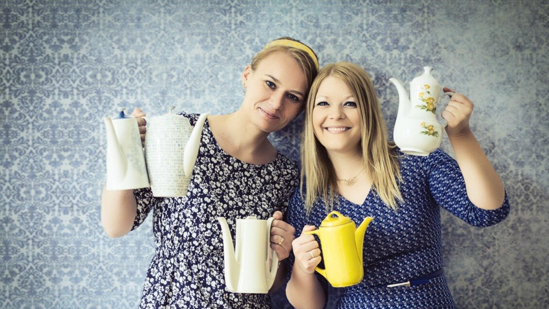 Zwei Frauen halten verschiedene Teekannen in den Händen und lächeln fröhlich. Der Hintergrund hat ein schönes, gemustertes Design. | © Omas Teekanne - Evgenia Rieger Photography