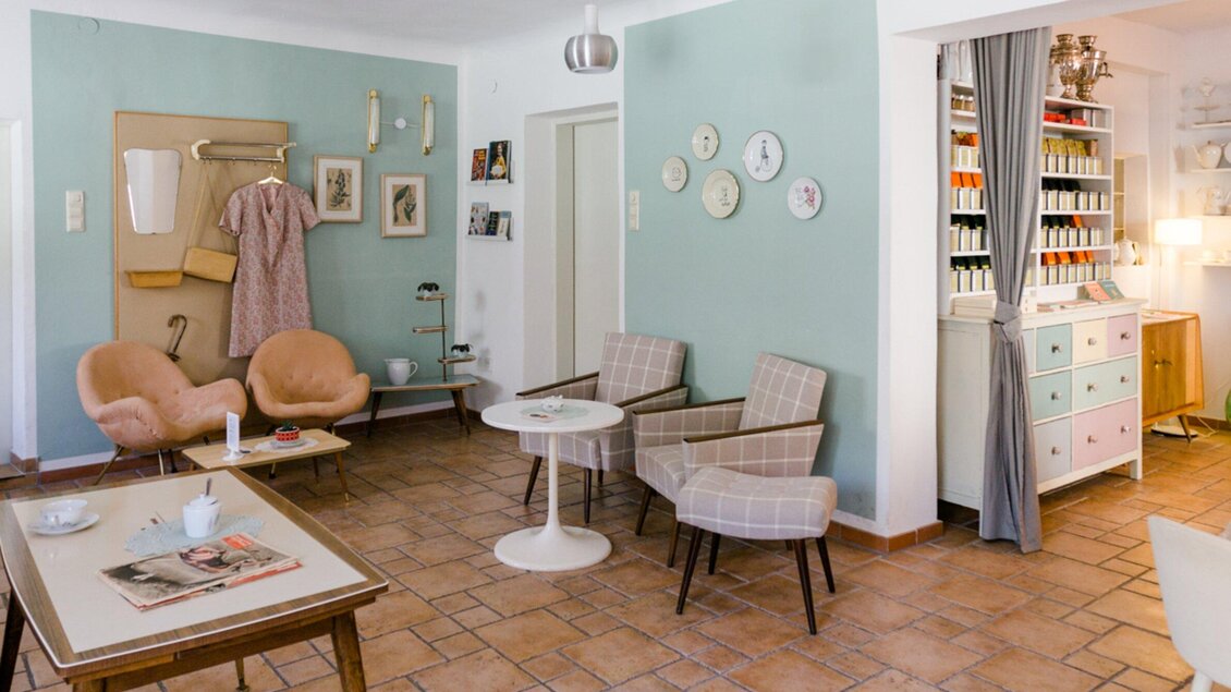 Ein stilvolles Wohnzimmer mit einem zentralen Tisch, bequemen Stühlen und einer freundlichen Farbgestaltung. An der Wand hängen verschiedene Bilder und eine Garderobe, die für ein einladendes Ambiente sorgt. | © Omas Teekanne