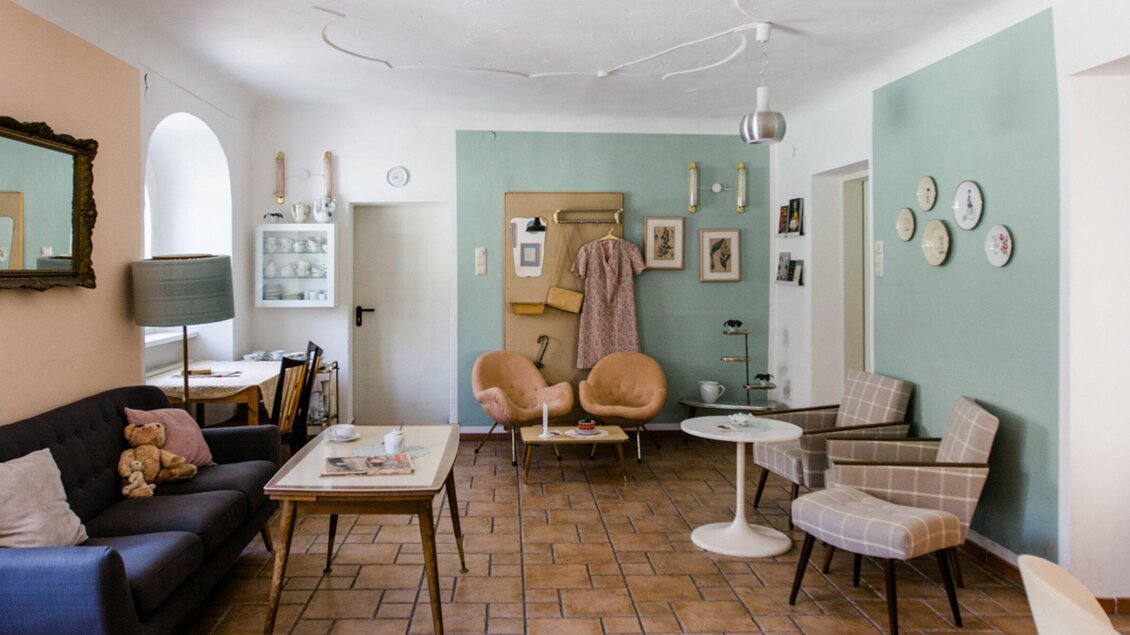 Ein freundliches Wohnzimmer mit einem blauen Sofa, Stilrichtungen aus der Mitte des Jahrhunderts und sanften Farben. Es gibt einen Tisch, gemütliche Sessel und dekorative Wanduhren. | © Omas Teekanne