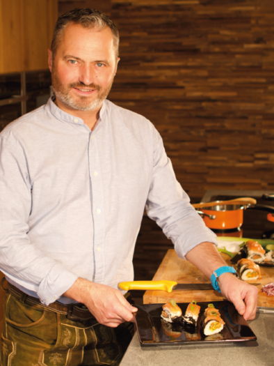 A chef stands in a modern kitchen and presents a plate of sushi. In the background, various cooking utensils and pots can be seen.
