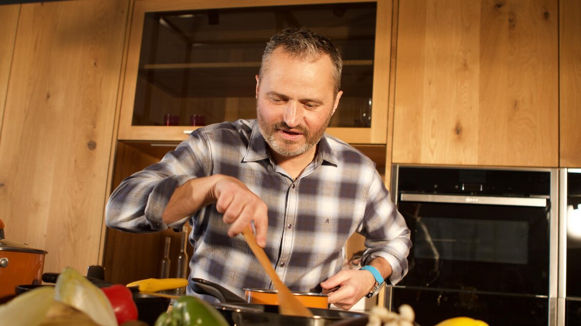 Ein Mann kocht in einer modernen Küche. Er rührt mit einem Holzlöffel in einem Topf, während frisches Gemüse auf dem Tisch liegt.