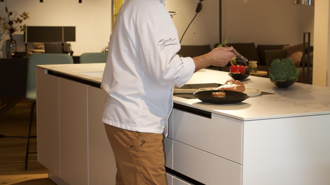 Ein Koch steht an einer modernen Kücheninsel und bereitet ein Gericht vor. Im Hintergrund sind stilvolle Möbel und eine Lichtquelle sichtbar.