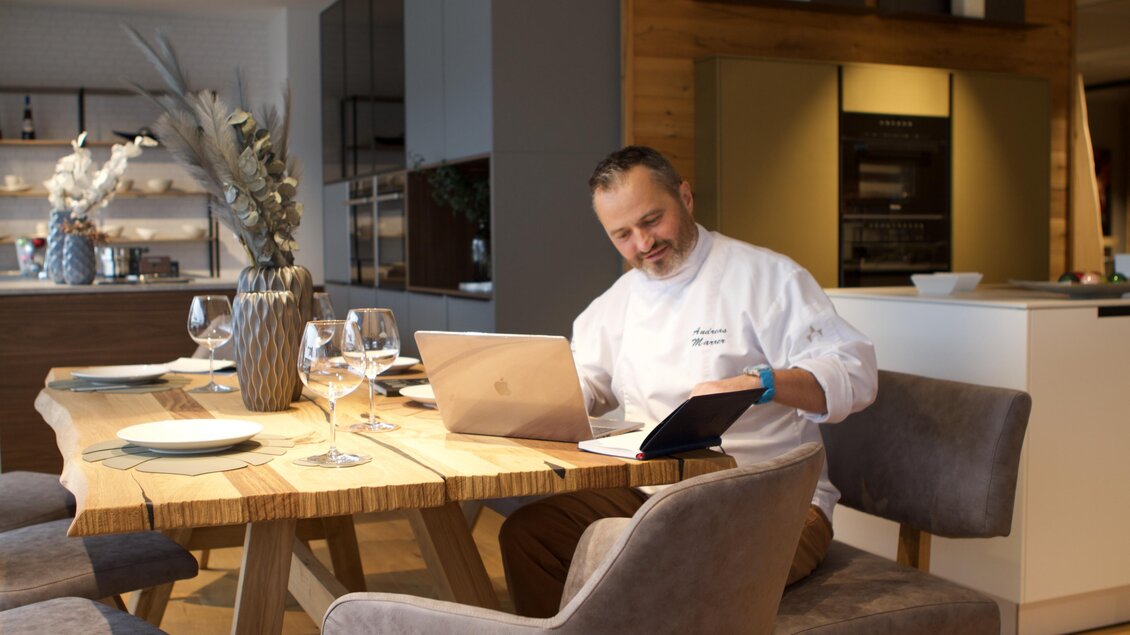 Ein Koch sitzt an einem Holztisch in einer modernen Küche und arbeitet an seinem Laptop. Um ihn herum sind gedeckte Teller und dekorative Elemente zu sehen.