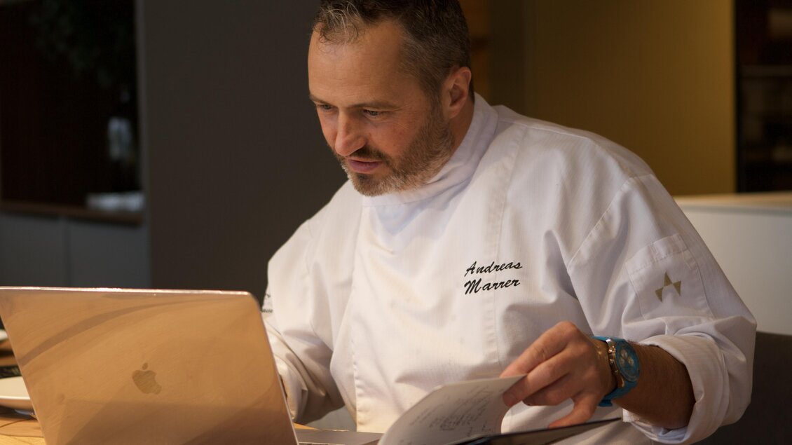 Ein Koch in weißer Berufskleidung arbeitet konzentriert an einem Laptop. Vor ihm liegt ein Kochbuch auf dem Tisch.