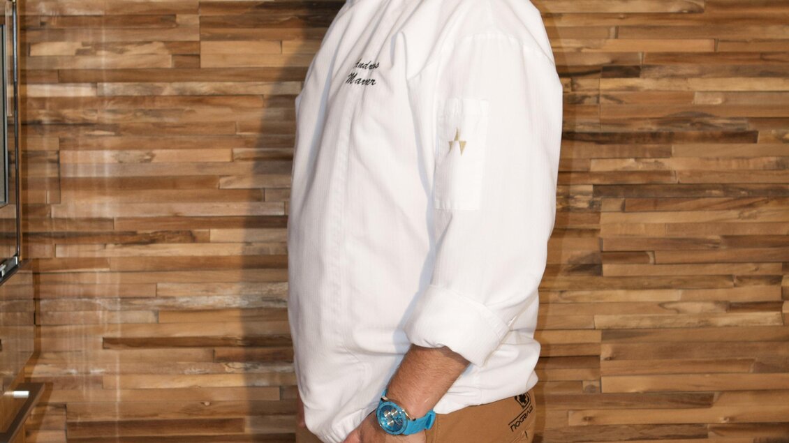 Ein lächelnder Koch steht seitlich vor einer Holzverkleidung. Er trägt eine weiße Kochjacke und braune Hose.