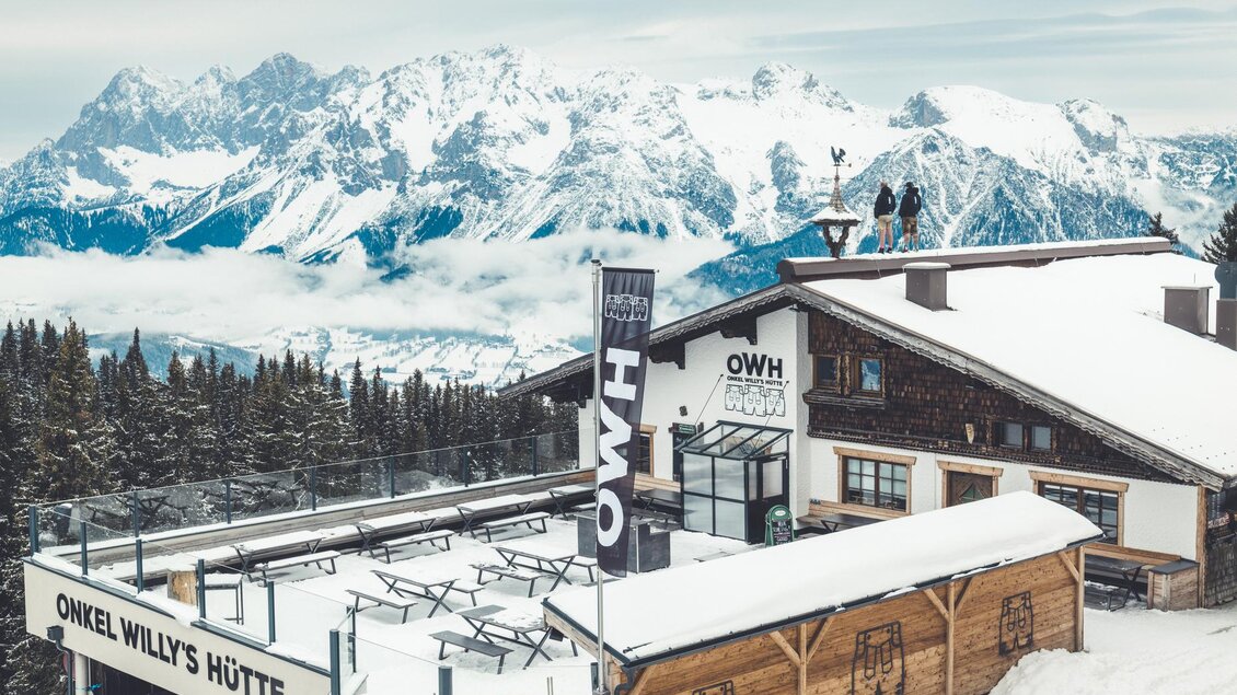 Eine Schneehütte in den Bergen mit schneebedeckten Gipfeln im Hintergrund. Der Himmel ist bewölkt und es gibt eine Terrasse mit Tischen. | © OWH