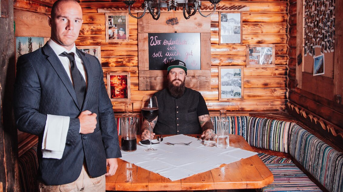 In einem gemütlichen Holzraum steht ein Mann in formeller Kleidung neben einem Tisch. Ein weiterer Mann sitzt am Tisch, umgeben von rustikalem Dekor und einer warmen Atmosphäre. | © OWH