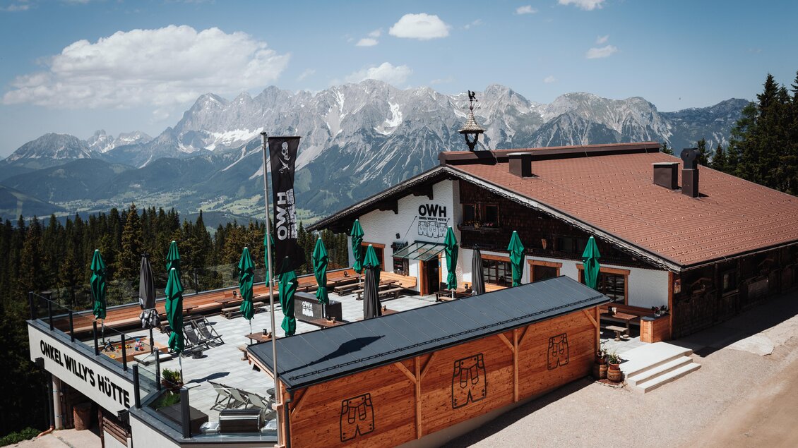 Ein Bergrestaurant mit einer großen Terrasse und grünen Sonnenschirmen. Im Hintergrund sind majestätische Berge und ein blauer Himmel zu sehen. | © OWH