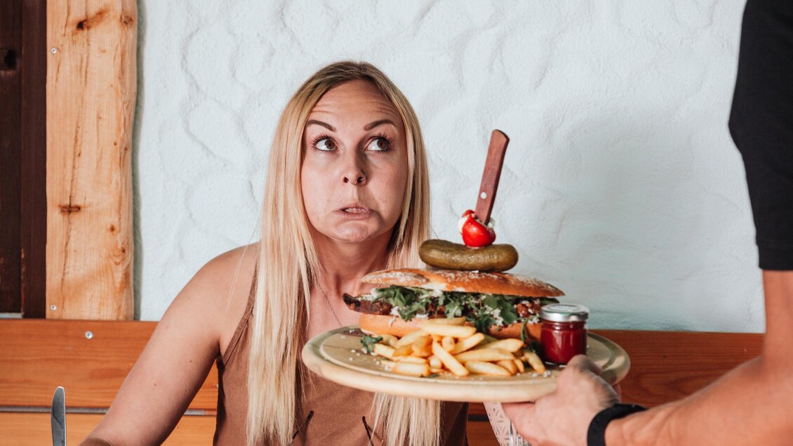 Eine überraschte Frau schaut auf einen Teller mit einem großen Burger und Pommes. Ein Kellner bringt das Essen und ein Bier an den Tisch. | © OWH
