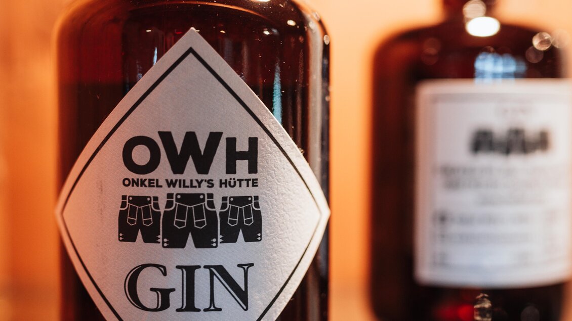Eine elegante Gin-Flasche mit einem auffälligen Etikett, das "Onkel Willy's Hütte" und "GIN" zeigt. Im Hintergrund ist eine weitere Flasche zu sehen. | © OWH