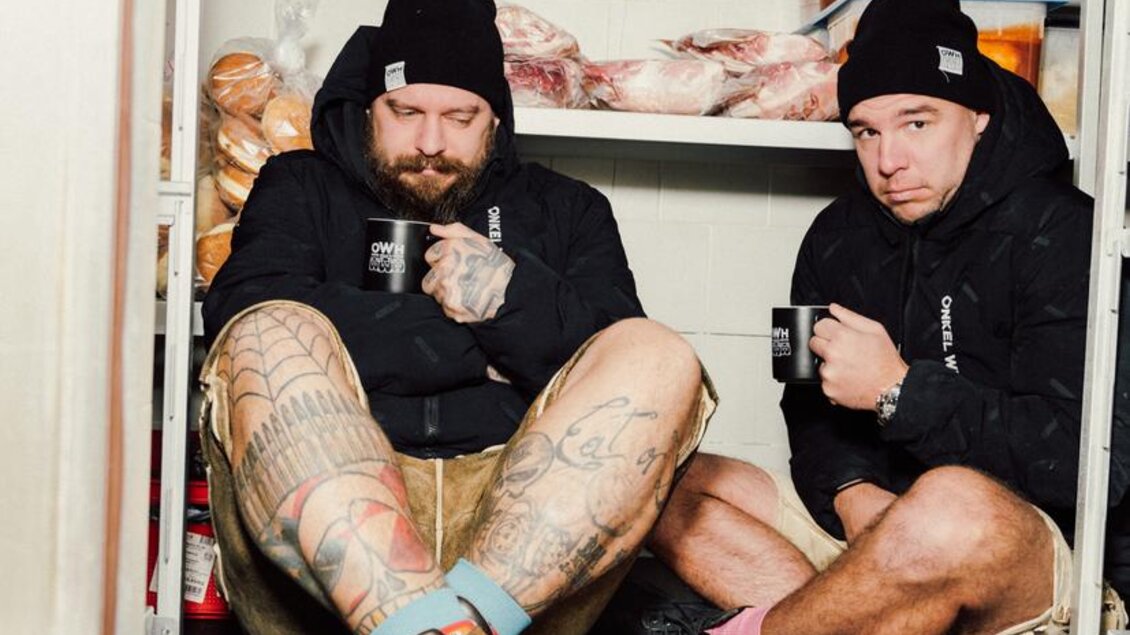 Zwei Männer sitzen in einem Kühlraum und schauen ernst. Der Raum ist mit Lebensmitteln gefüllt, und sie tragen bequeme Kleidung sowie Mützen. | © OWH