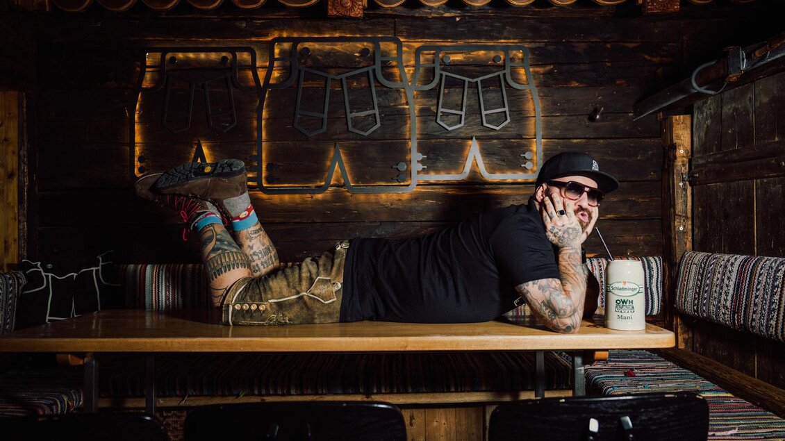 Ein bärtiger Mann in Sonnenbrille liegt lässig auf einem Holztisch. Im Hintergrund sind dekorative Elemente und eine bunte Bank zu sehen. | © OWH