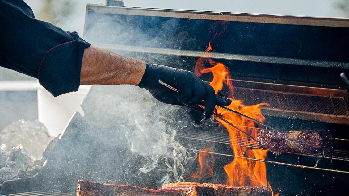 Ein Grill mit Flammen und rauchenden Fleischstücken. Eine Person mit Handschuhen wendet das Grillgut. | © OWH