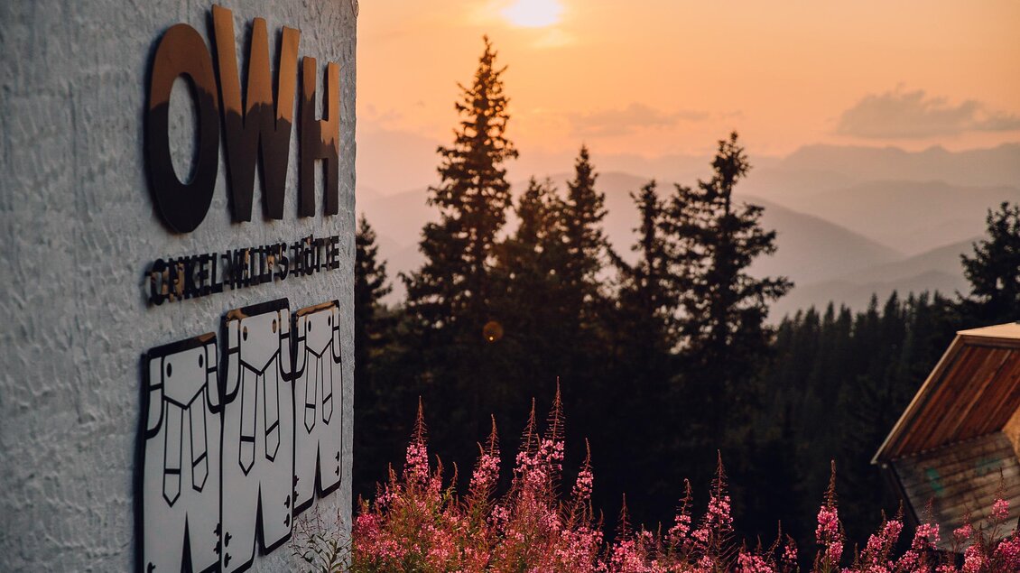 Ein malerischer Sonnenuntergang über Bergen, mit einer blühenden Wiese im Vordergrund. Im Bild ist das Logo einer Lodge zu sehen. | © OWH