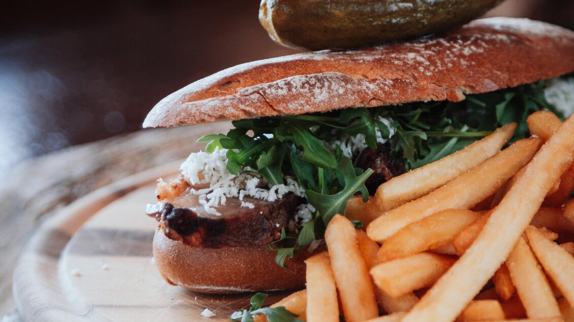 Ein leckerer Burger mit frischer Salatbeilage, garniert mit einer Gurke und einer Tomate auf einem Holzbrett. Dazu gibt es knusprige Pommes frites. | © OWH