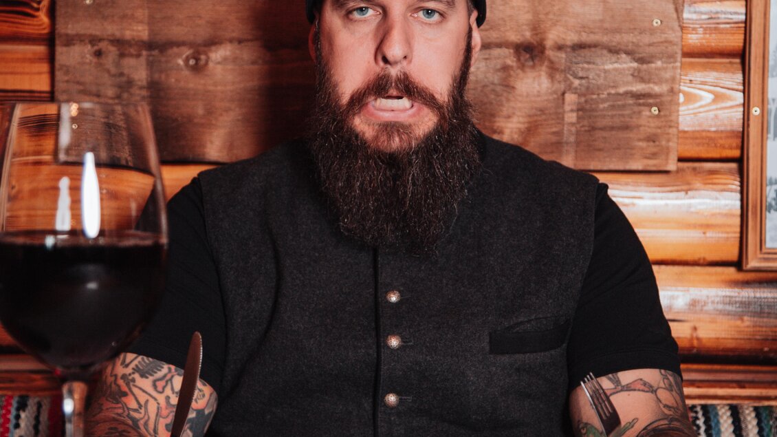 Ein Mann mit Bart sitzt an einem Tisch in einem rustikalen Restaurant und schaut ernst. Vor ihm liegt ein Teller mit Essen und ein großes Glas Rotwein. | © OWH