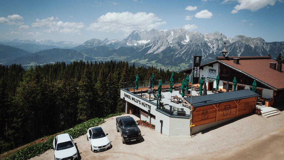 Eine Berghütte mit Terrassenstühlen und schönen Ausblick auf die Berge. Im Vordergrund stehen mehrere Autos auf einem geschotterten Parkplatz. | © OWH