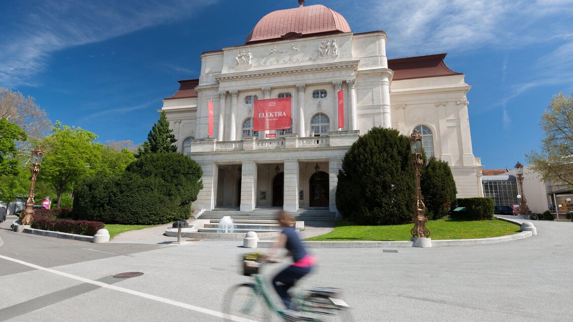 Die Grazer Oper mit einer markanten Kuppel und einem roten Banner. Im Vordergrund fährt eine Person auf einem Fahrrad vorbei. | © Graz Tourismus - Harry Schiffer
