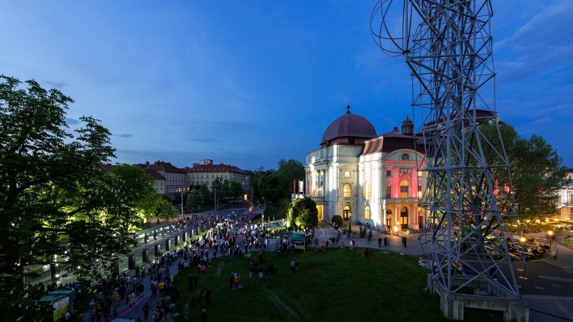 Die festlich beleuchtete Oper Graz am Abend mit vielen Menschen im Freien. Umgeben von Bäumen und eine Stahlbau Skulptur im Hintergrund. | © Graz Tourismus - Harry Schiffer