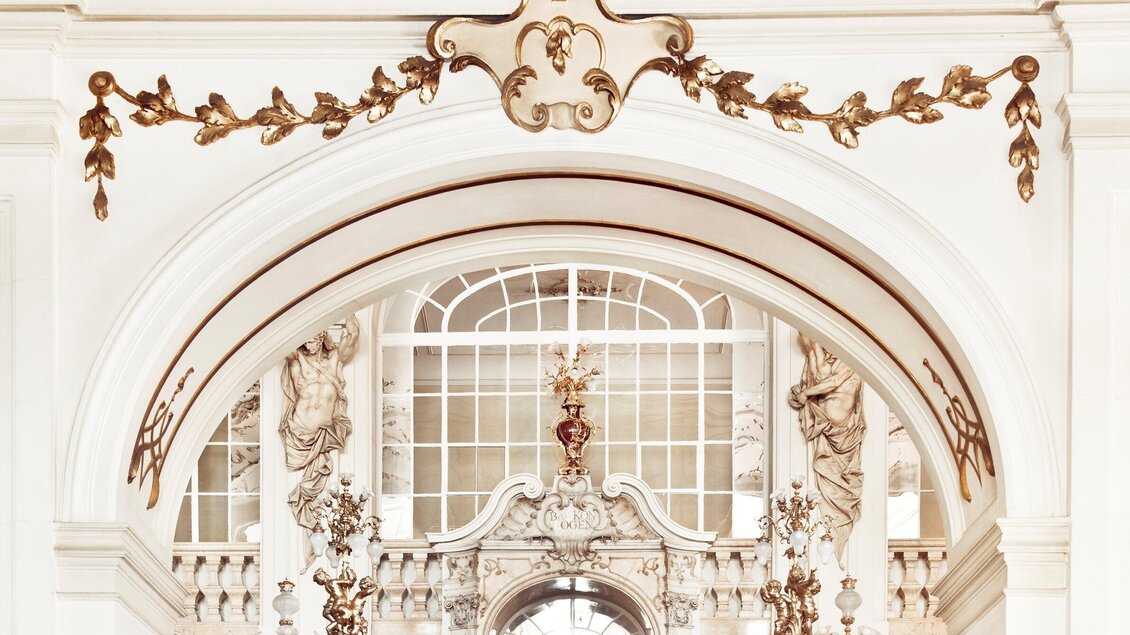 Ein prächtiger Stukbogen mit goldenen Verzierungen und kunstvollen Details in der Oper Graz. Im Hintergrund sind große Fenster, die viel Licht einlassen. | © Opernhaus Graz +