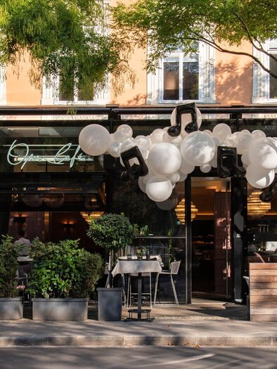 A modern restaurant with a glass facade and an inviting entrance area. In front of the restaurant, there are plants and outdoor tables. | © Operncafé - Jauk Katharina