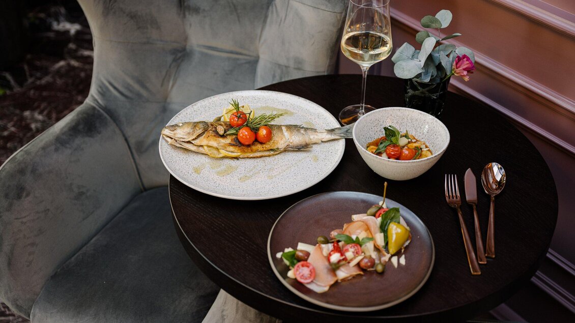 Ein elegantes Tischarrangement mit gegrilltem Fisch, frischen Tomaten und einer bunten Salatbeilage. Dazu ein Glas Wein und Besteck auf einem dunklen Tisch. | © Operncafé - Joel Kernasenko