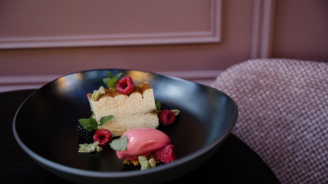 Ein köstliches Dessert auf einem schwarzen Teller. Es besteht aus einer Torte, Himbeeren und einem pinken Sorbet. | © Operncafé - Joel Kernasenko