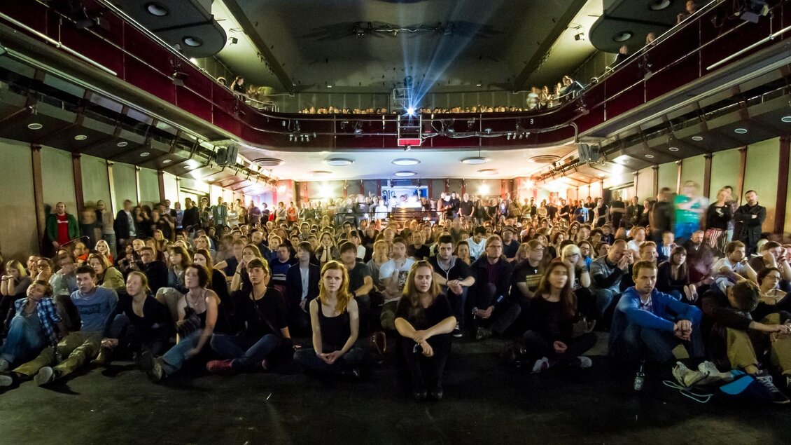 Ein großes Publikum sitzt im Saal und schaut gespannt nach vorne. Die Beleuchtung sorgt für eine lebendige Atmosphäre. | © Julien Duval