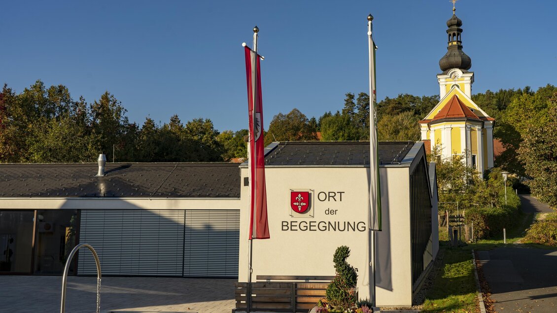 Ein modernes Gebäude mit dem Schriftzug „Ort der Begegnung“ im Vordergrund. Im Hintergrund ist eine Kirche mit einer auffälligen Türe und einem Turm zu sehen. | © Kurkommission Bad Blumau