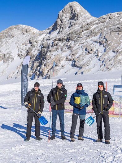 A group of four people stands in a snow-covered landscape with mountains in the background. They are wearing winter clothing and holding shovels in their hands. | © René Eduard Perhab