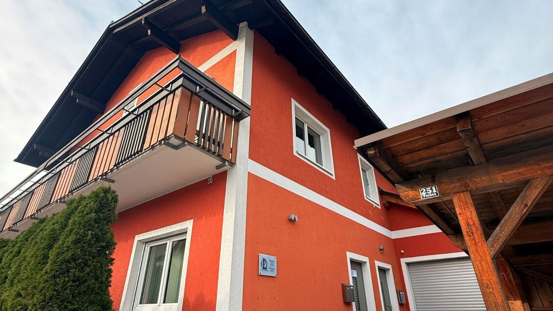 Ein modernes Gebäude in kräftigem Orange mit einem Balkon und großen Fenstern. Im Vordergrund sind einige Bäume und eine Holzstruktur sichtbar. | © Lisa Schweitzer