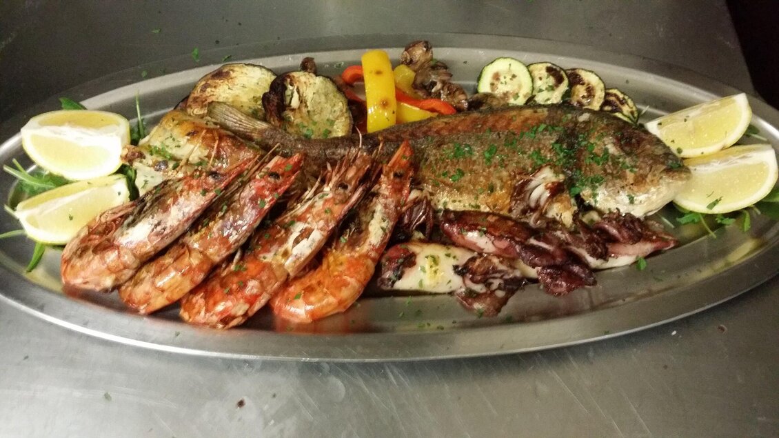 Ein platter mit frischem Fisch und Meeresfrüchten, darunter Garnelen und Tintenfisch. Dazu liegen Zitronenscheiben und bunte Gemüse. | © Osteria dei Sensi