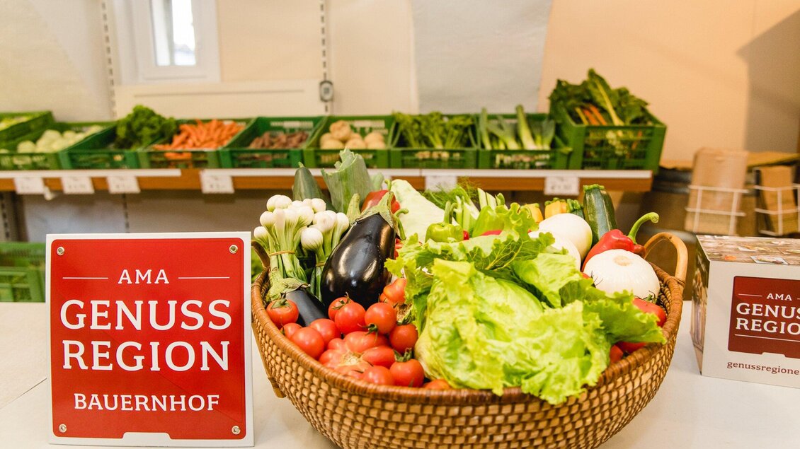Ein Korb mit frischem Gemüse steht auf einem Tisch. Im Hintergrund sind grüne Kisten mit weiteren Gemüseprodukten zu sehen. | © AMA GENUSS REGION - Mias Photoart