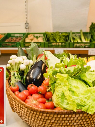 Ein Korb mit frischem Gemüse steht auf einem Tisch. Im Hintergrund sind grüne Kisten mit weiteren Gemüseprodukten zu sehen. | © AMA GENUSS REGION - Mias Photoart