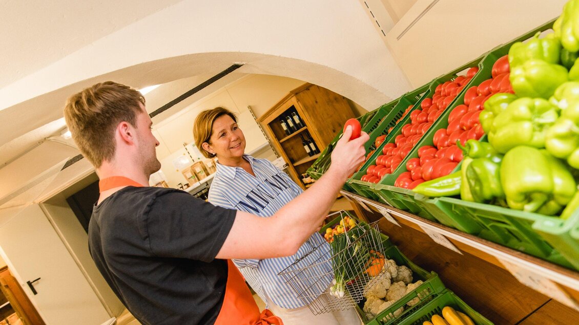 Ein Verkäufer zeigt frisches Gemüse in einem Lebensmittelgeschäft. Eine Kundin schaut interessiert zu und lächelt. | © AMA GENUSS REGION - Mias Photoart