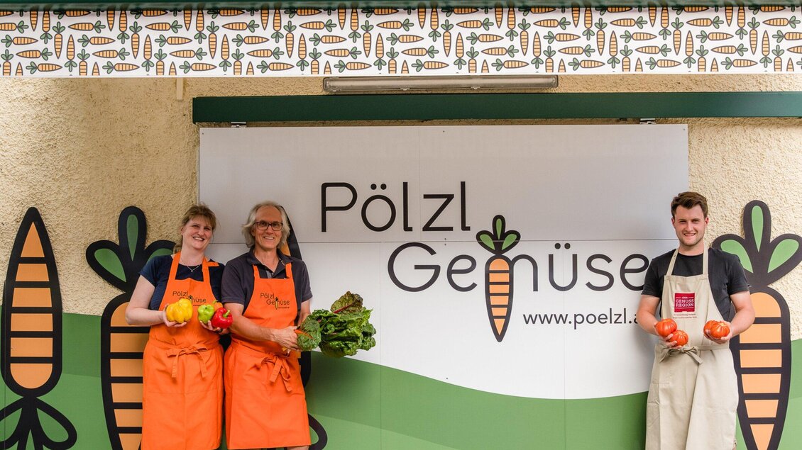 Drei Personen stehen vor einem Banner mit dem Schriftzug "Pölzl Gemüse". Sie halten frisches Gemüse in den Händen und tragen orangefarbene Schürzen. | © AMA GENUSS REGION - Mias Photoart