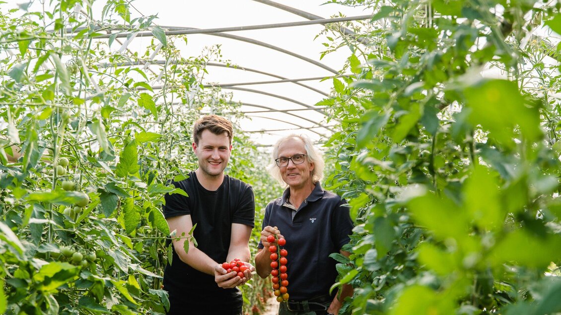 Zwei Männer stehen in einem Gewächshaus zwischen grünen Pflanzen und halten frische Tomaten in den Händen. Sie lächeln und sehen glücklich aus. | © AMA GENUSS REGION - Mias Photoart
