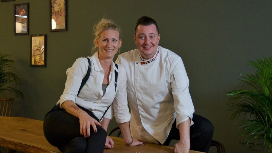 Die Besitzer Eva und Hannes Michetschläger in Service- und Kochkleidung sitzen lächelnd auf einem Holztisch. | © PEP