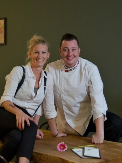 The owners Eva and Hannes Michetschläger in service and cooking attire sit smiling at a wooden table. | © PEP