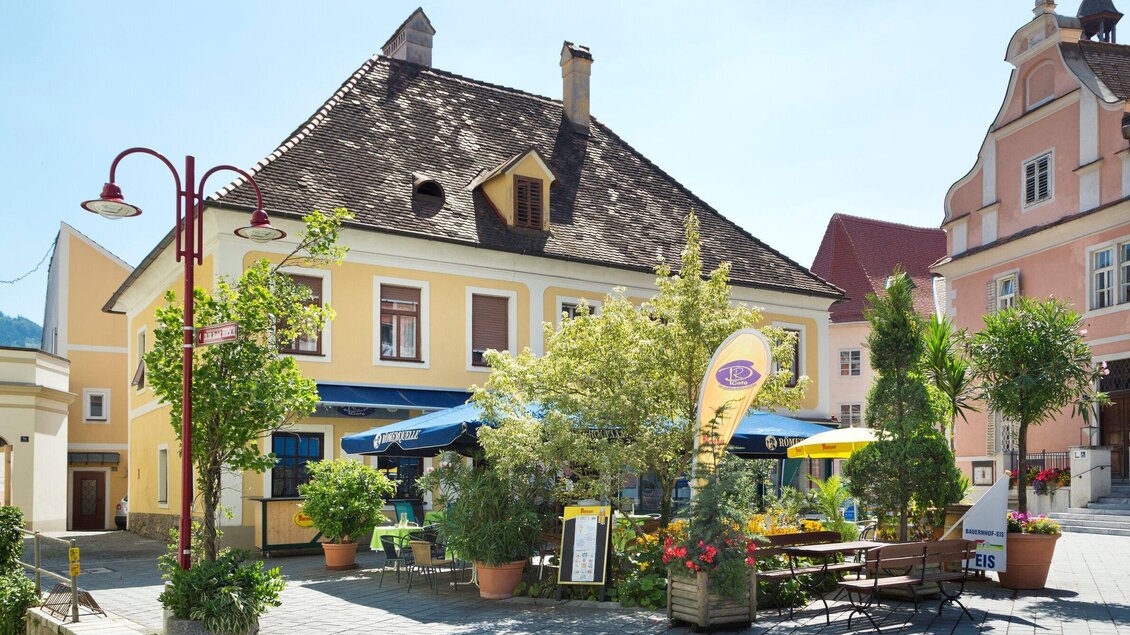 Ein charmantes Restaurant in einer sonnigen Straße, umgeben von bunten Pflanzen und Terrassenmöbeln. Die gelbe Fassade des Gebäudes strahlt Gemütlichkeit aus. | © TV Region Graz - René Vidalli