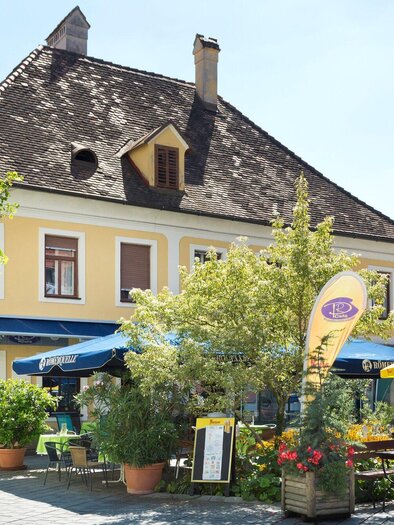 Ein charmantes Restaurant in einer sonnigen Straße, umgeben von bunten Pflanzen und Terrassenmöbeln. Die gelbe Fassade des Gebäudes strahlt Gemütlichkeit aus. | © TV Region Graz - René Vidalli