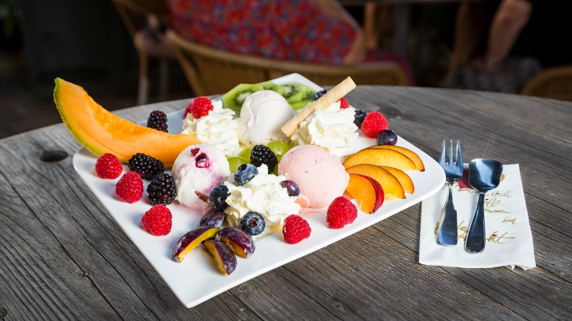 Ein köstlicher Eisbecher mit verschiedenen Eissorten, frischen Früchten und Sahne. Serviert auf einem weißen Teller auf einem Holztisch. | © TV Region Graz - René Vidalli