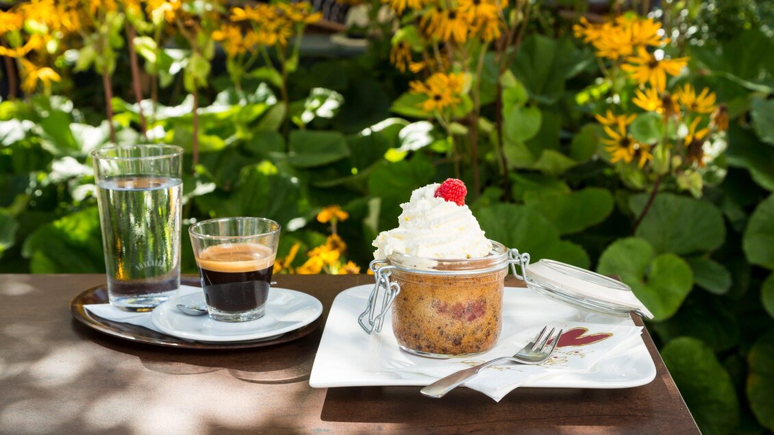 Ein hübsches Dessert in einem Glas mit Sahne und einer Himbeere, serviert auf einem Teller. Daneben stehen ein Espresso und ein Glas Wasser auf einem Tisch in einer blühenden Umgebung. | © TV Region Graz - René Vidalli