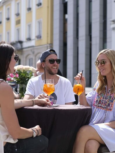 Drei Freunde sitzen im Freien an einem Tisch und genießen ihre Getränke. Umgeben von bunten Blumen und sonnigem Wetter strahlt die Szene eine entspannte Atmosphäre aus. | © Michael Maili